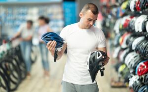 A man in a white t-shirt compares two bicycle helmets inside a bike shop, surrounded by rows of helmets on display.