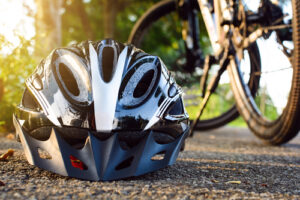 Close-up of a black bicycle helmet on the ground next to a parked bike, with sunlight filtering through trees in the background.