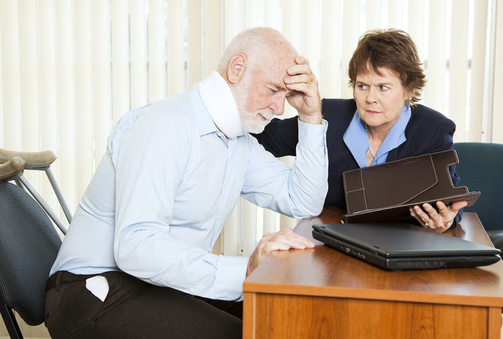 An older man with a neck brace looking distressed with a concerned woman holding a tablet.