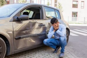 A man in jeans and a blue shirt covers his face with his hands, upset next to his damaged car with dents and scratches after an accident.