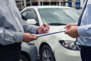 An insurance agent inspects a damaged car while a customer signs a claim form after a traffic accident. Concept of insurance claims.