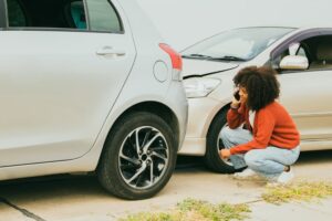 Shocked African American woman involved in a car accident, sits near the damaged vehicle, calling insurance for assistance.