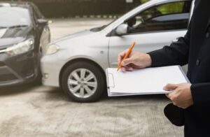 Insurance officer notes details on clipboard as agent inspects black car post-accident.