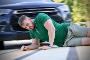 A man in a green shirt and shorts lies on the ground in pain after being hit by a car, with the front of the vehicle visible in the background, highlighting the severity of pedestrian accidents.