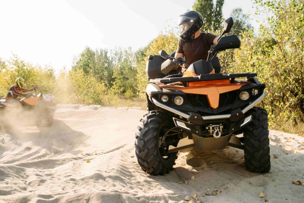 Two people riding all-terrain vehicles on a sandy trail through the woods, wearing helmets for off-road safety.