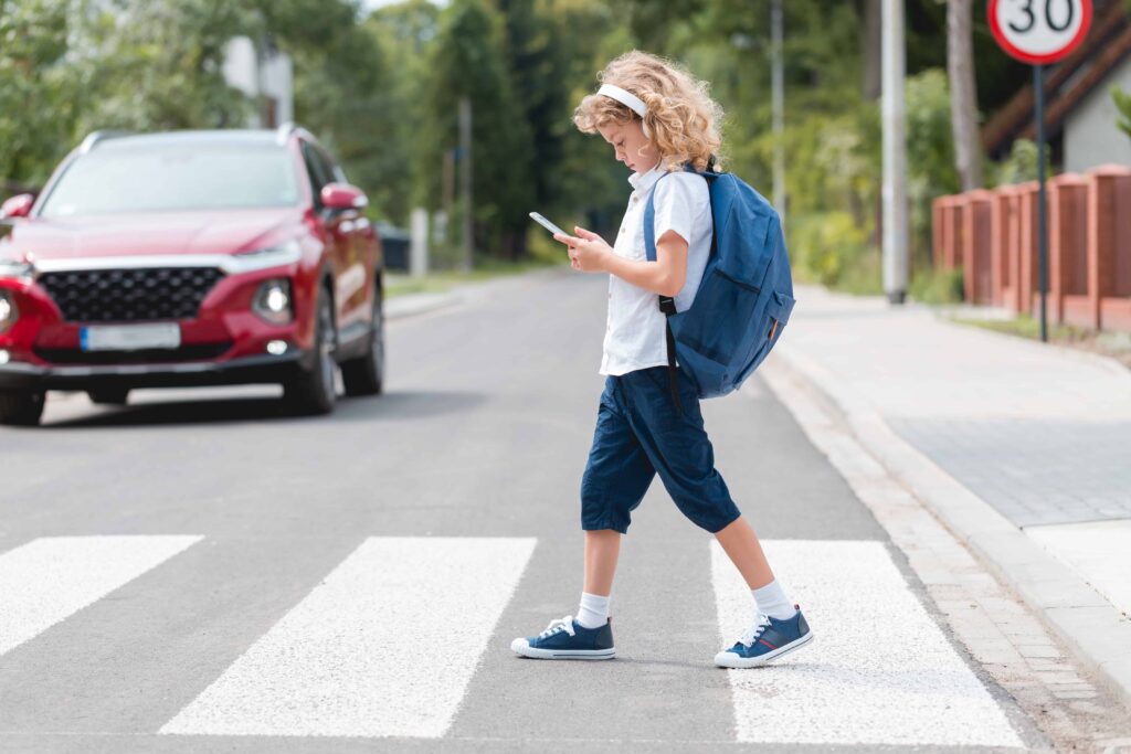 Child with backpack looking at phone while crossing street on crosswalk with approaching car.