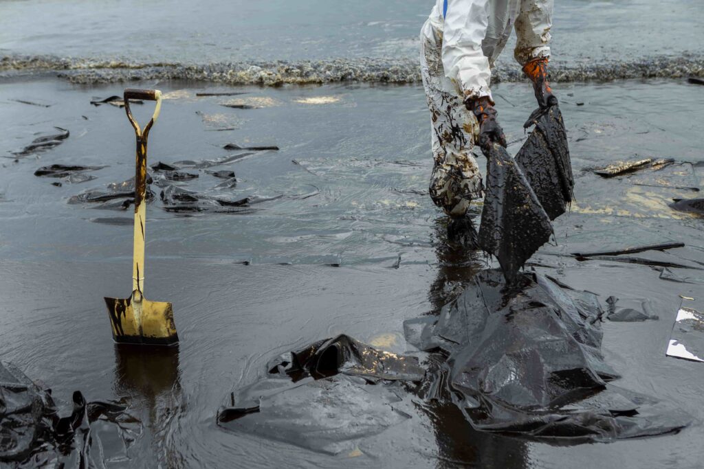 Worker in protective suit cleaning oil spill on beach with shovel and absorbent materials.